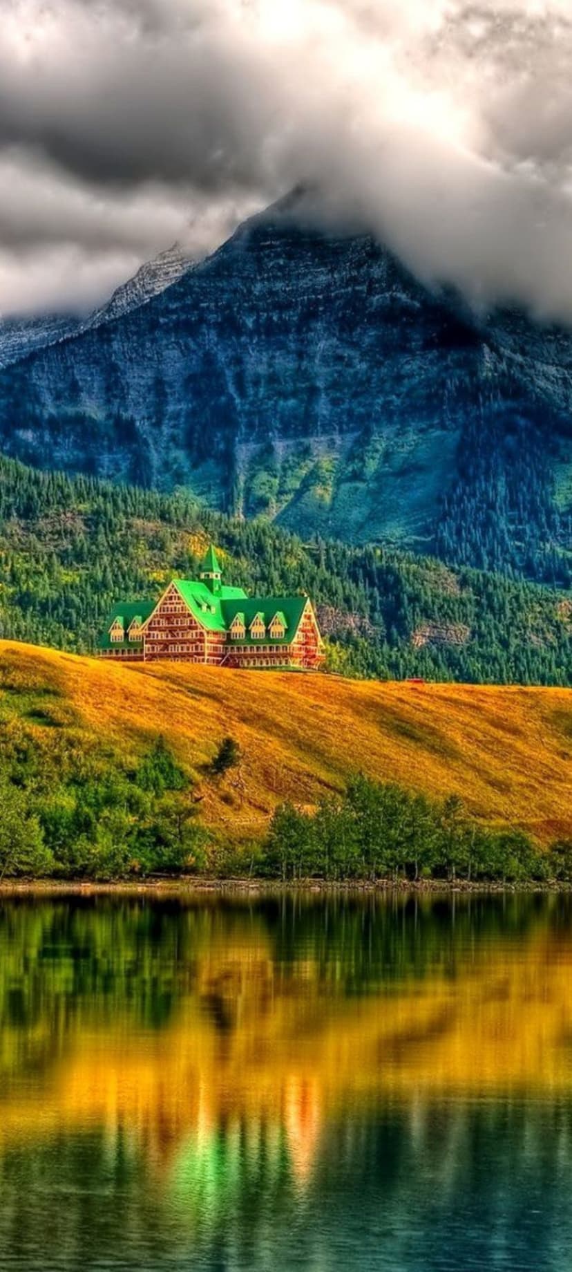 Grand hotel with a green roof reflects in a still blue lake surrounded by golden hills and misty mountains.