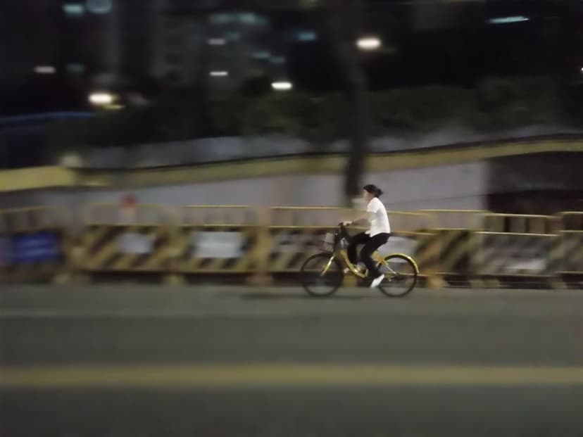 Cyclist in a white top rides a yellow bike past streetlights with motion blur on a dark city road at night.