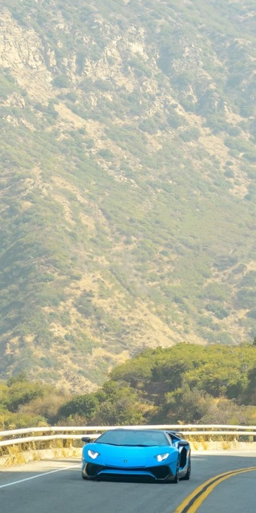 Blue Lamborghini Aventador supercar speeds along a curved mountain road with lush green cliffs in the background.