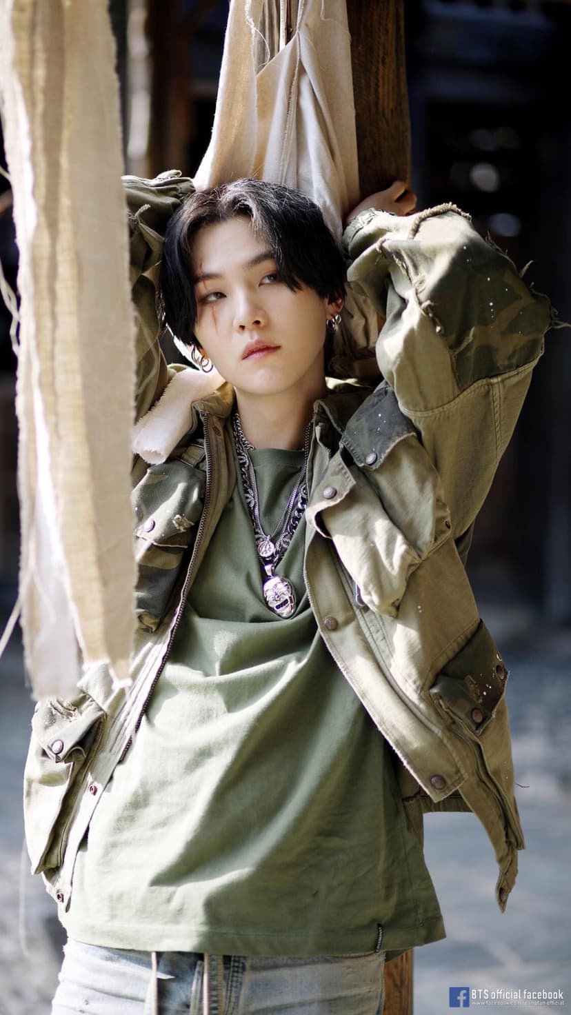 Suga from BTS in an olive green jacket and silver necklaces poses against a weathered, gritty background.