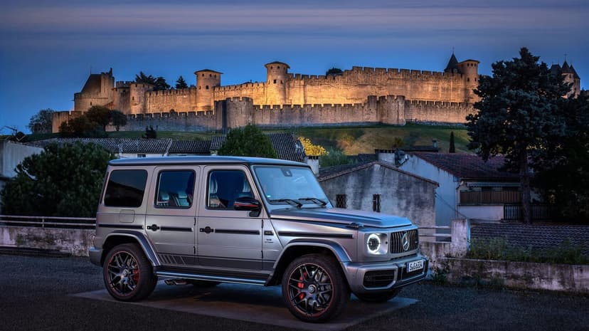 Silver Mercedes-Benz G63 AMG SUV parked in front of a lit medieval fortress under a deep blue twilight sky.