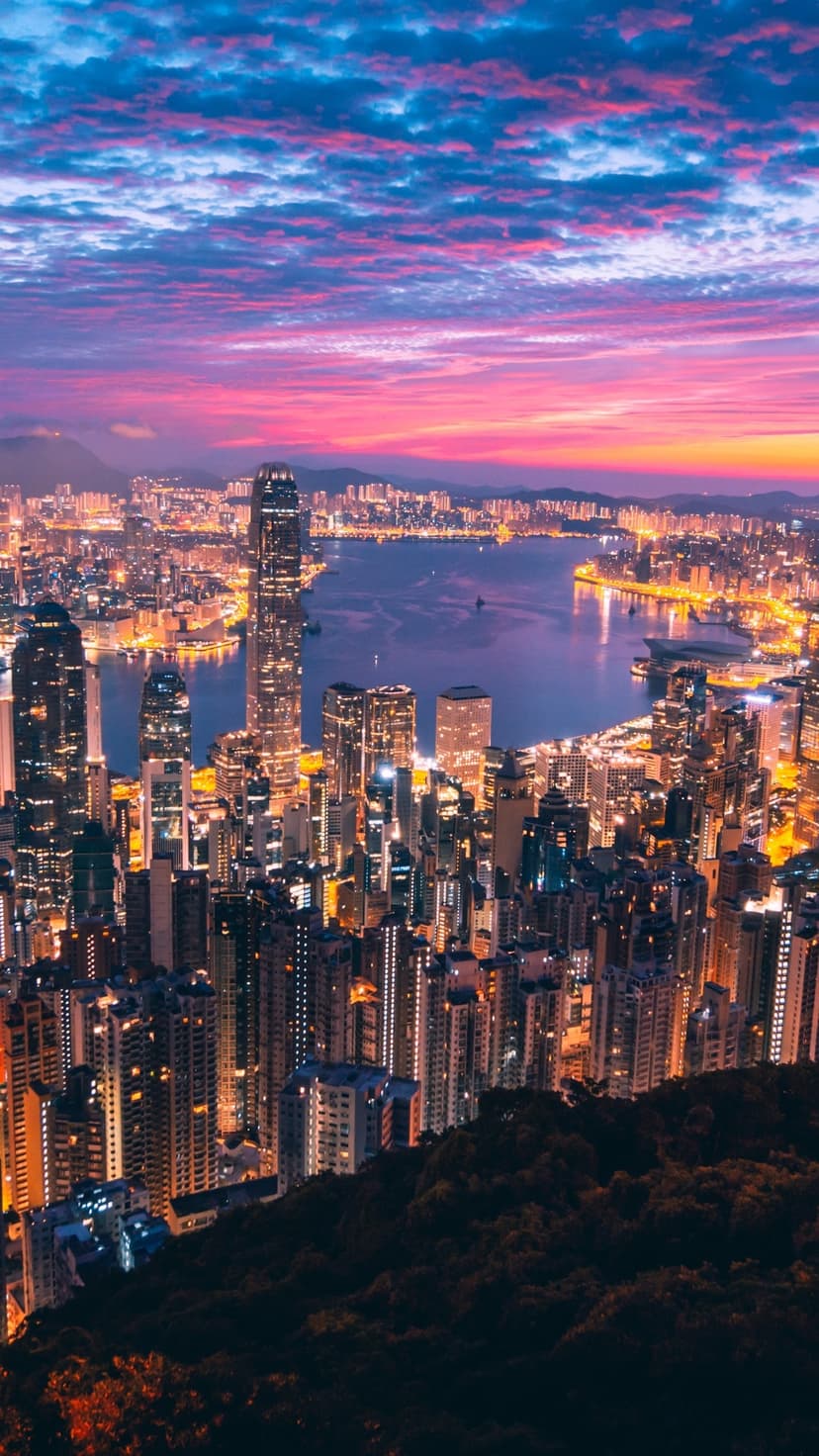 Illuminated skyscrapers border a coastal harbor under a purple and orange twilight sky from an aerial view.