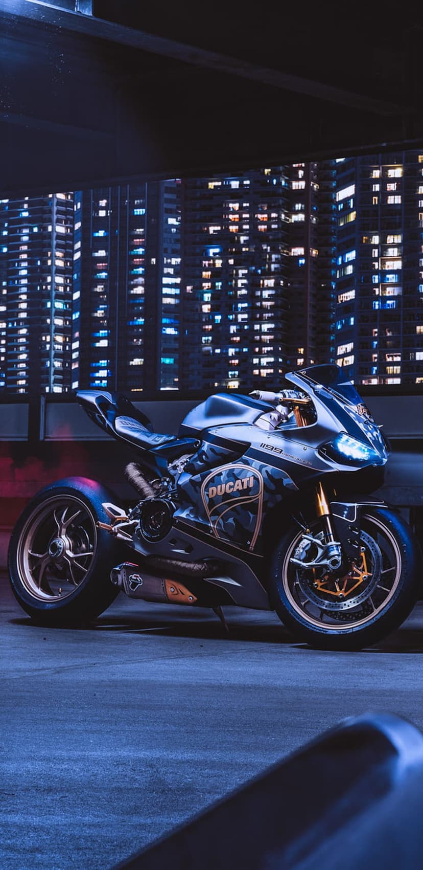 Ducati sportbike with bronze accents parked on a city street at night under blue lights and bokeh skyscrapers.