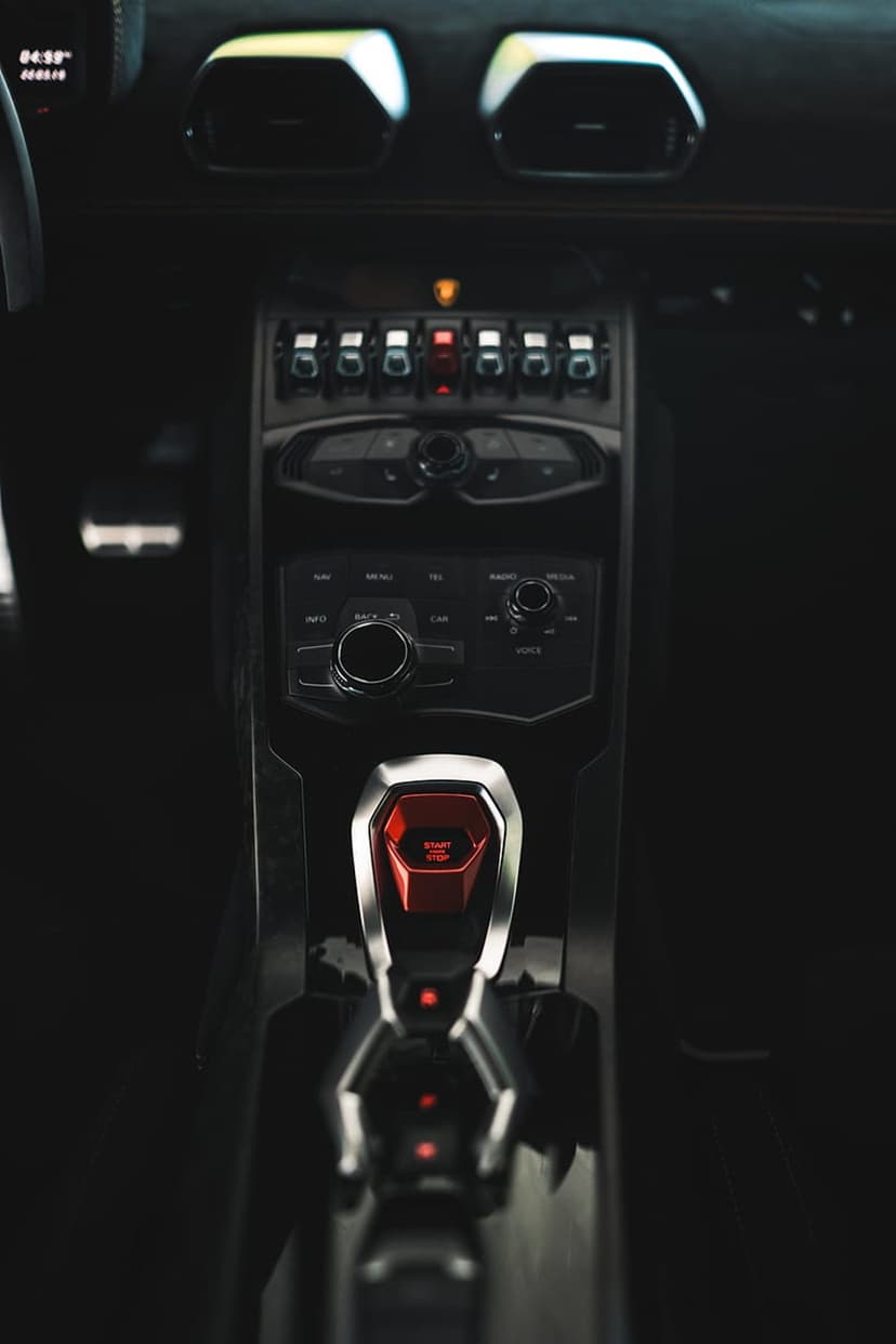 Red engine start button with a flip cover sits amidst the carbon fiber console and vents of a supercar cockpit.