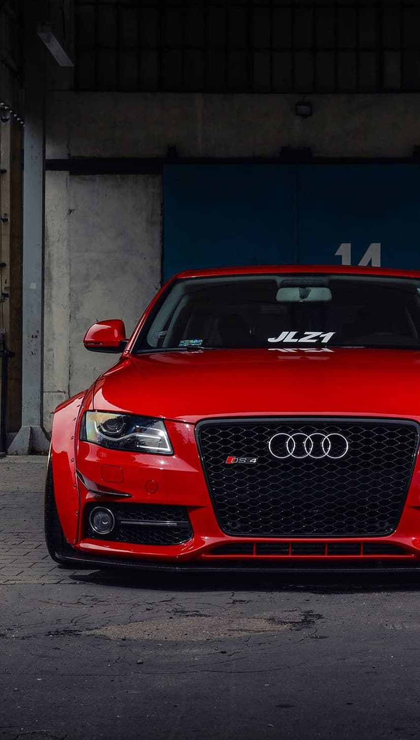 Red Audi RS4 featuring widebody fenders and a custom bumper parked in front of a blue industrial garage door.