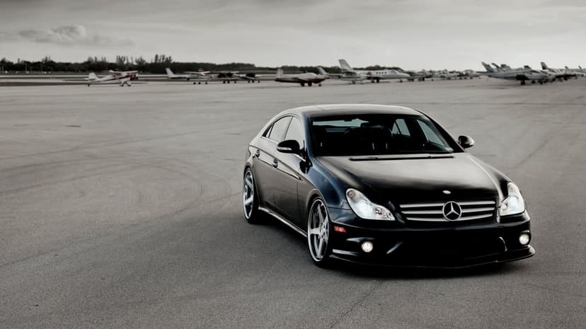 Black Mercedes CLS sedan with silver wheels parked on an airport tarmac with small planes under a cloudy sky.