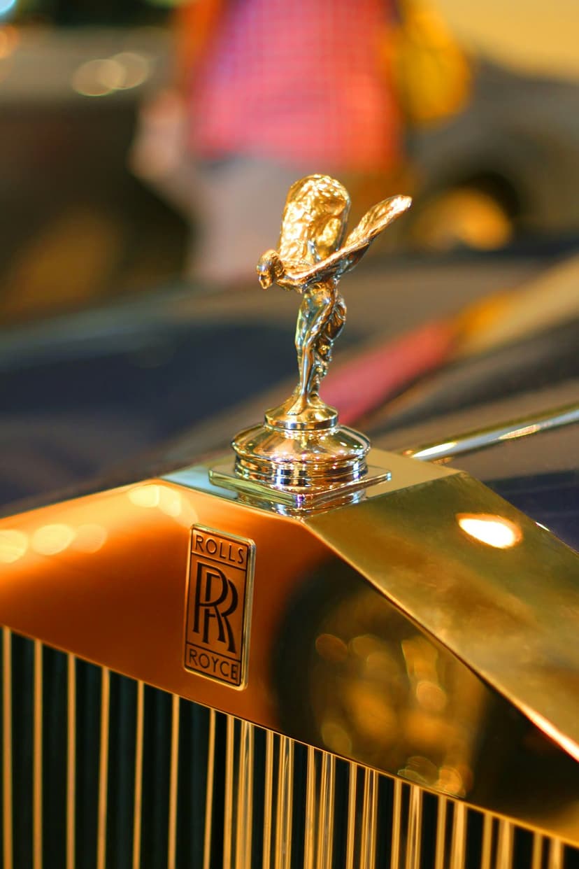 Spirit of Ecstasy statuette in polished gold perched on a Rolls-Royce emblem with a metallic car grille below.