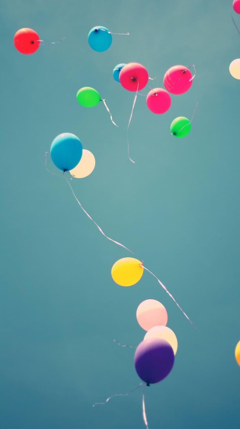 Vibrant red, blue, and yellow balloons with trailing ribbons float upward against a bright, cloudless blue sky.