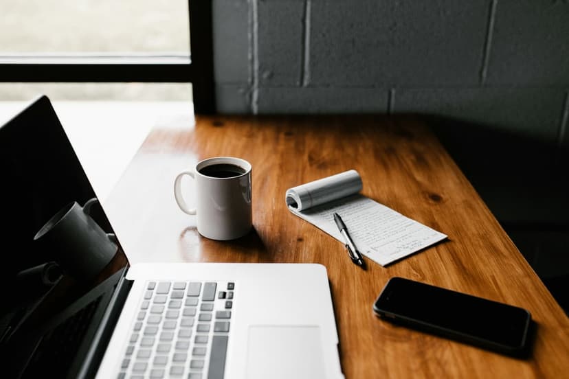 Laptop, steaming coffee mug, and smartphone rest on a light wood grain desk illuminated by natural sunlight.