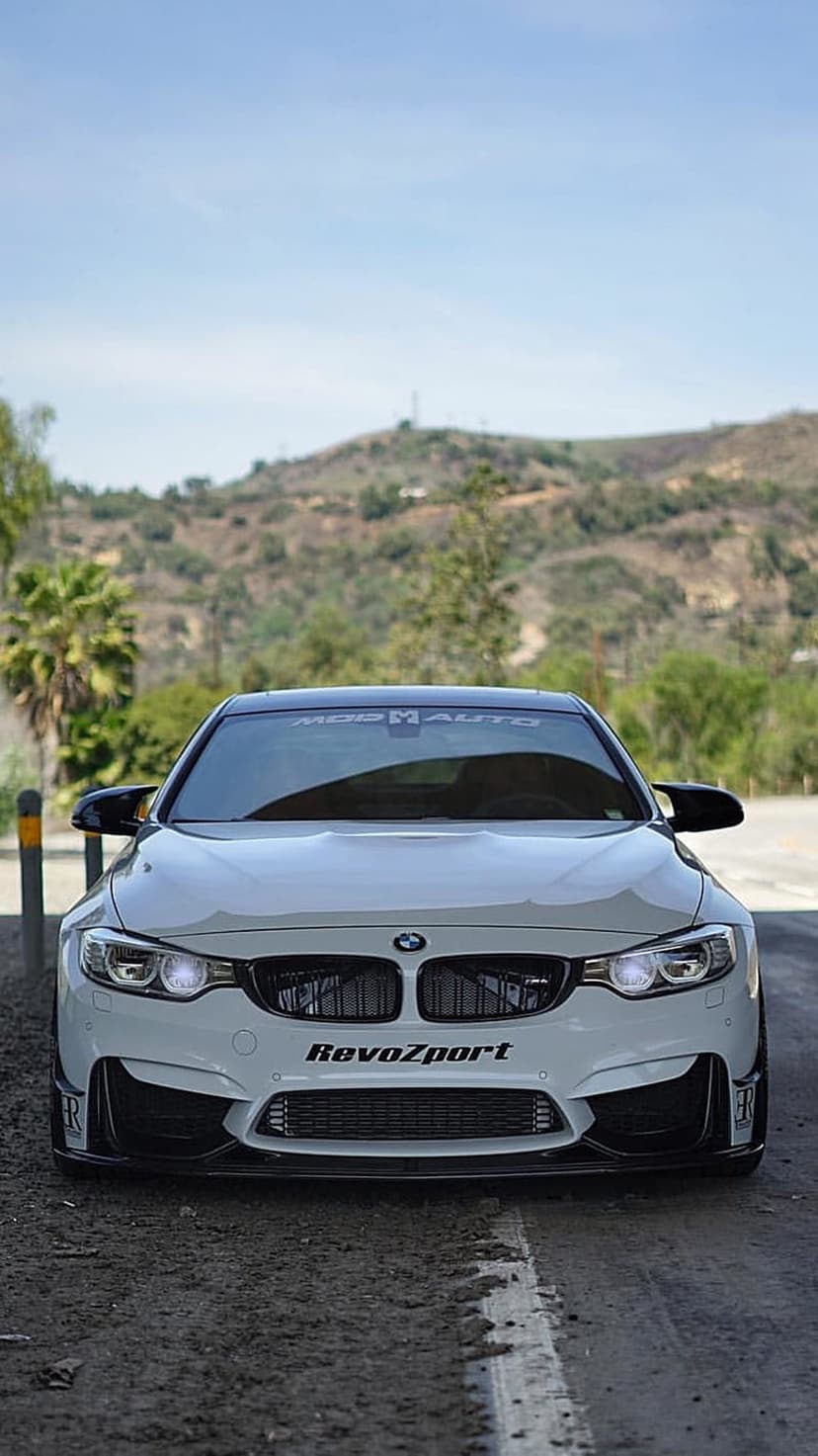 White BMW M4 front view with RevoZport kit parked on a paved road against green hills and a blue sky.