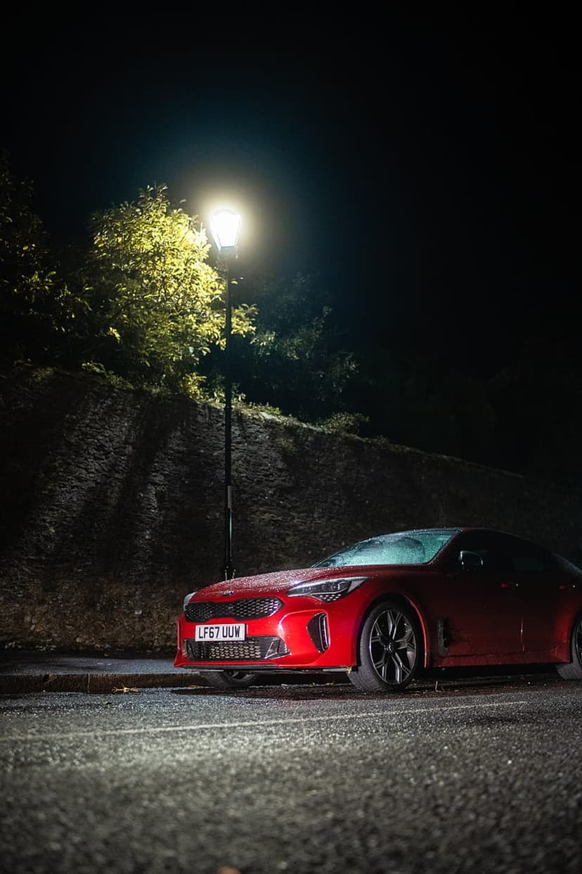 Red Kia Stinger parked on wet asphalt beneath a bright street lamp at night with dark foliage nearby.