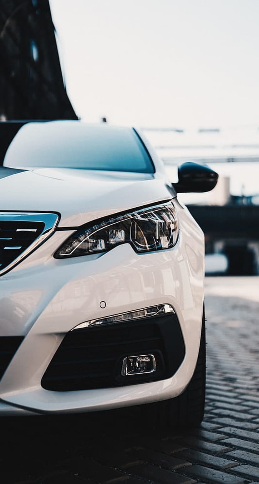 White Peugeot 308 front grille and LED headlights against a blurred cobblestone street and bright blue sky.