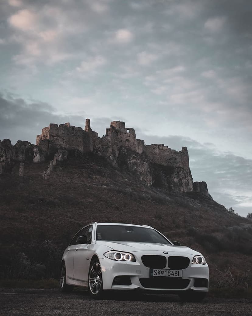 White BMW station wagon parked before crumbling stone castle ruins under a dramatic, moody overcast sky.