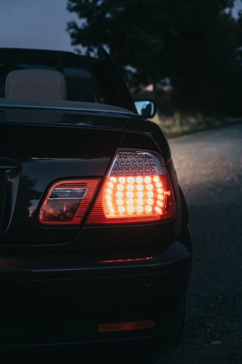 Red LED taillight of a dark convertible car illuminated at dusk on a quiet asphalt road lined with trees.