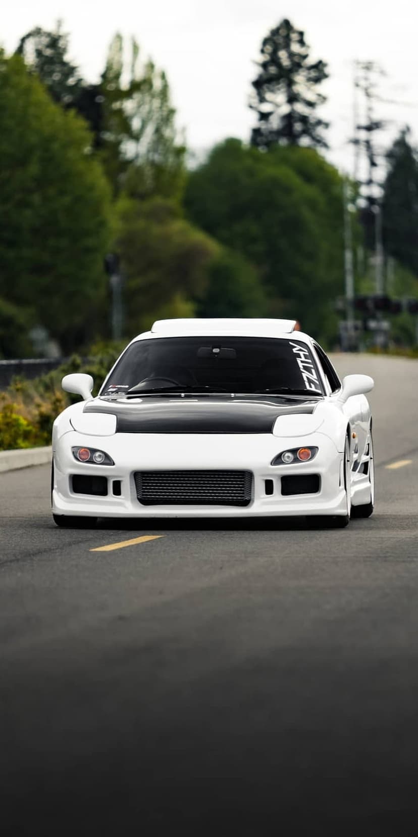 White Mazda RX-7 sports car with a black hood drives on an asphalt road surrounded by green trees.