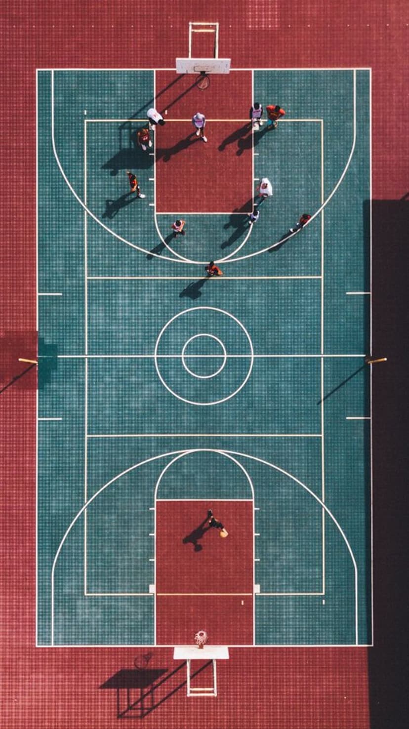 Athletes play basketball on a red and teal outdoor court viewed from a direct top-down aerial perspective.
