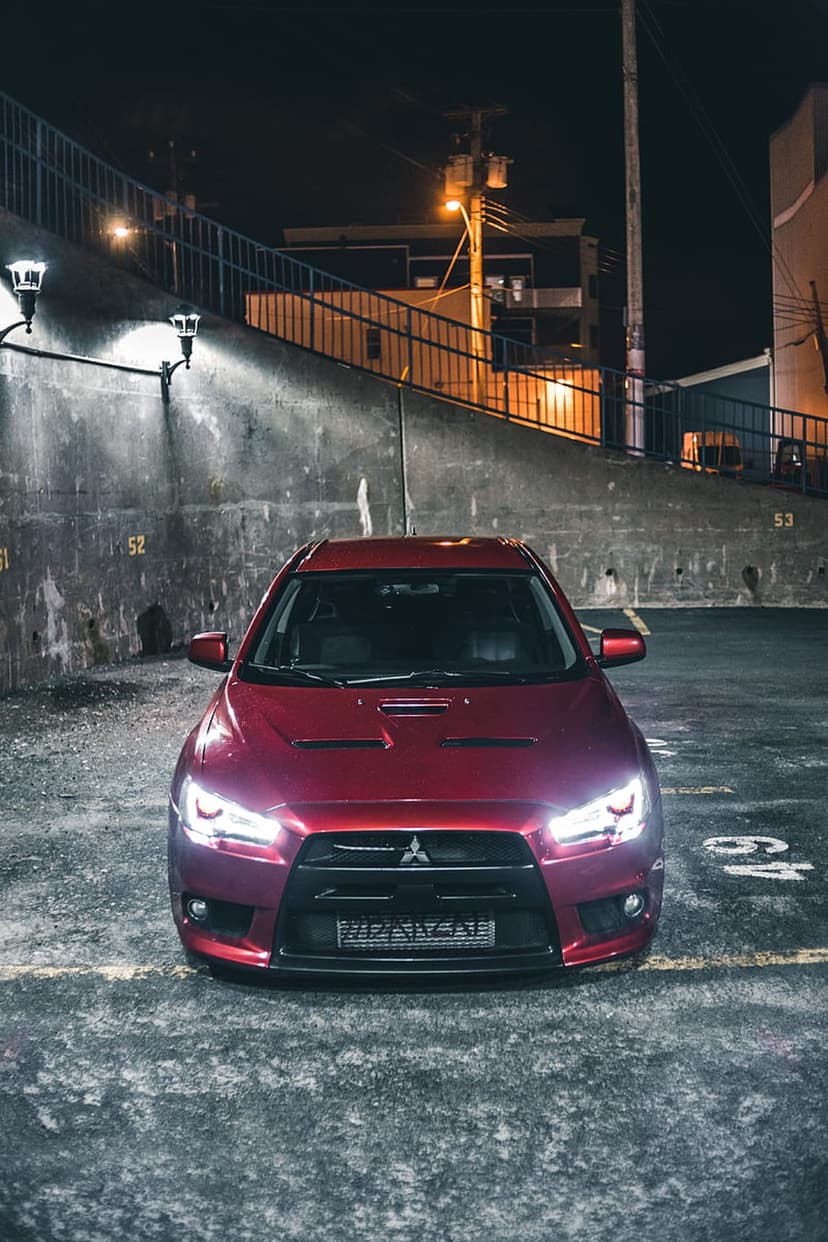 Red Mitsubishi Lancer Evolution with glowing headlights parked in a dark urban industrial lot at night.