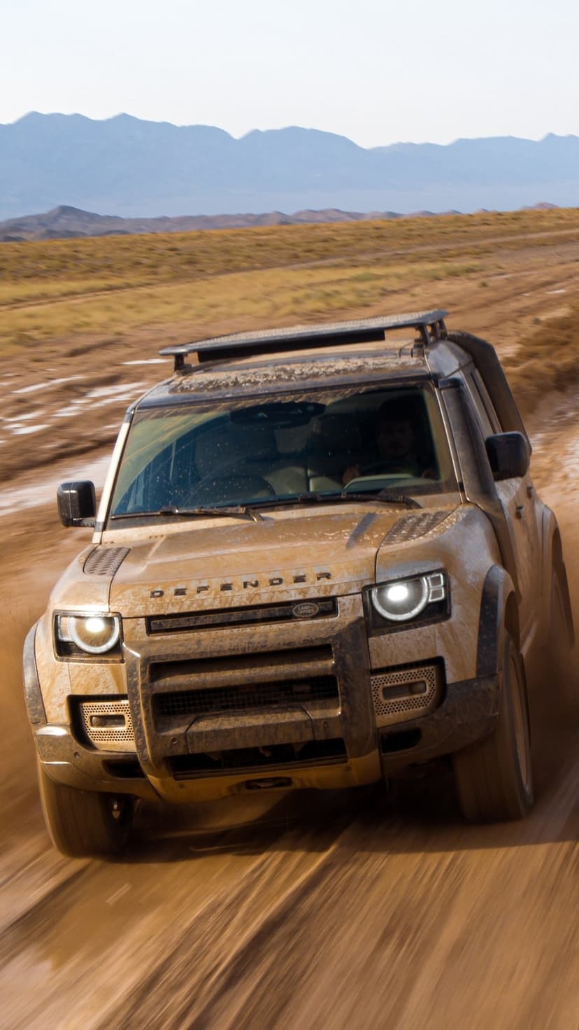 Muddy Land Rover Defender driving fast on a dirt road with mountains in the background under clear sky.