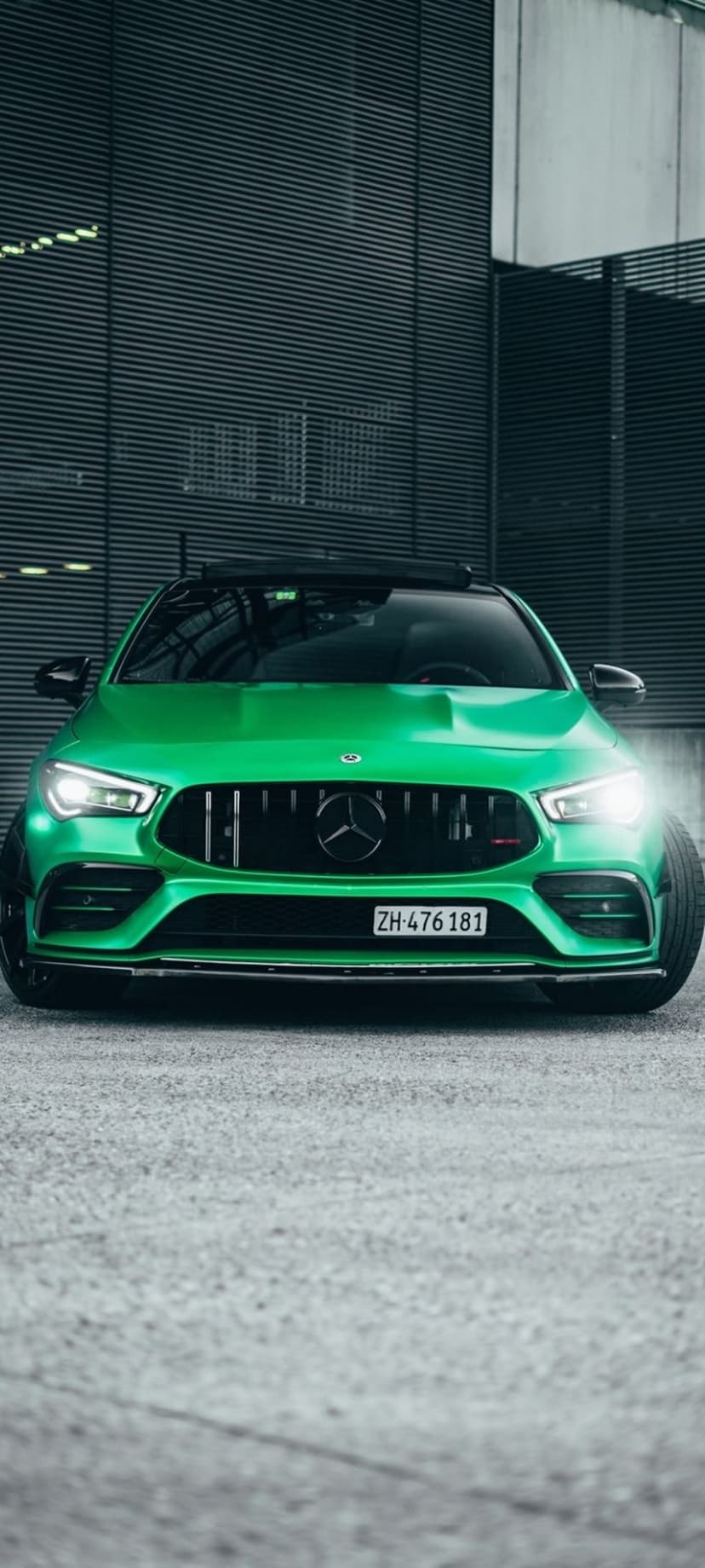 Green Mercedes-AMG CLA 45 S sports car with a vertical-slat grille parked in front of a modern gray building.