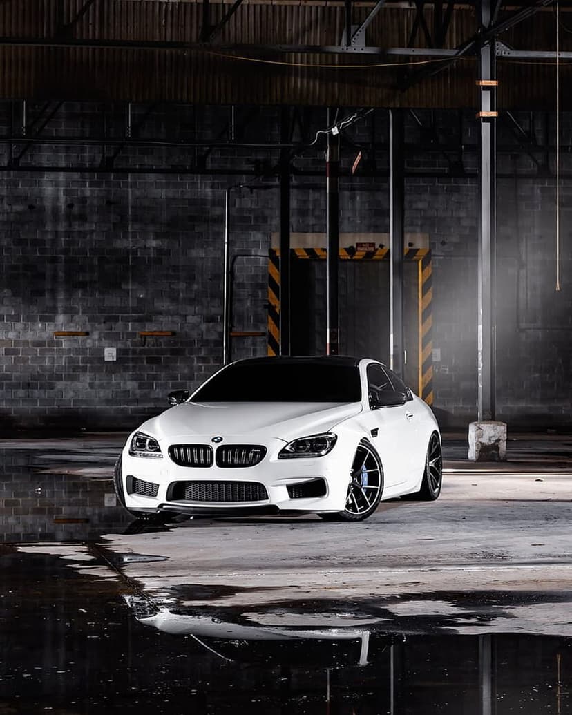 White BMW M6 Coupe parked on a wet garage floor with dramatic overhead lighting and dark brick walls behind it.