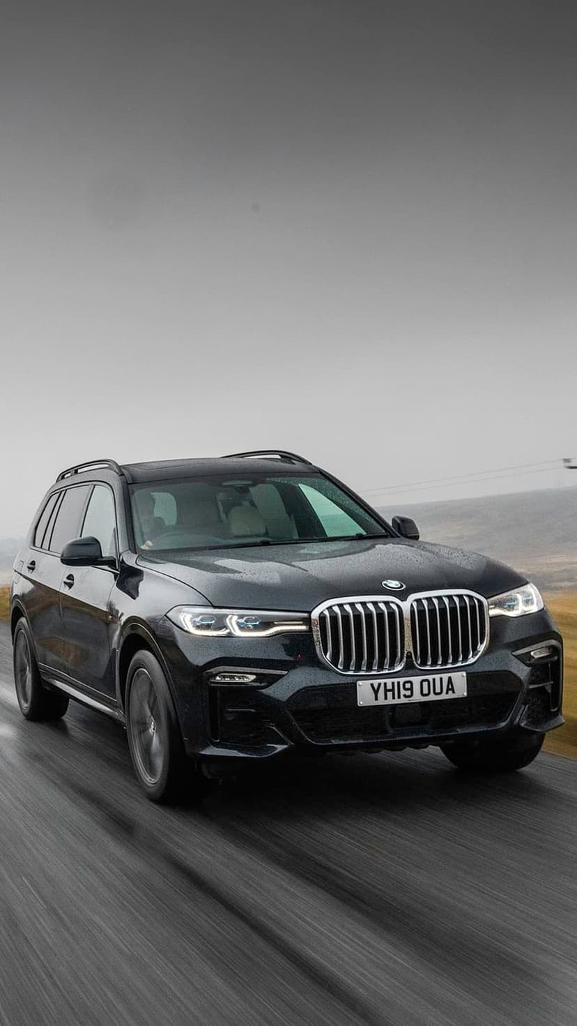 Black BMW X7 SUV driving on a wet asphalt road with bright headlights reflecting off the damp surface.
