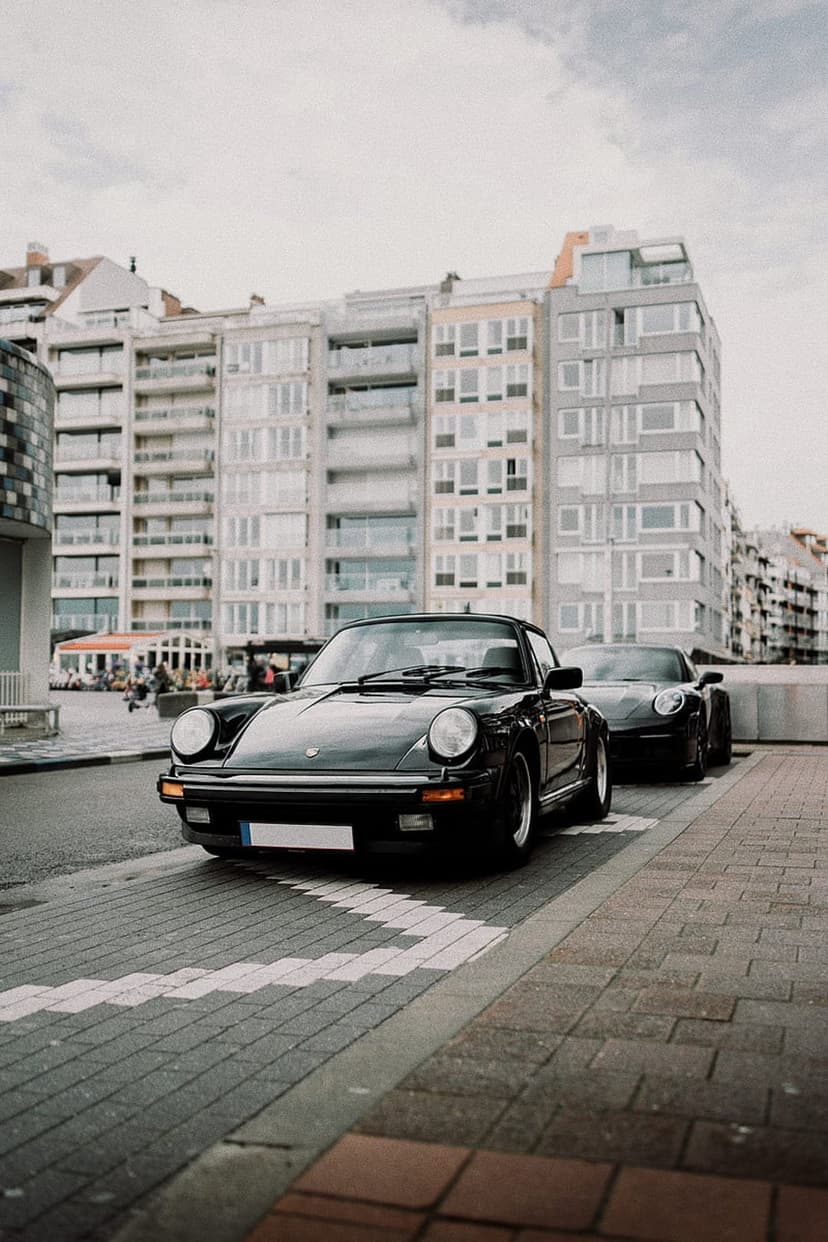 Pair of black Porsche 911 classic cars parked on cobblestones with modern waterfront architecture behind them.