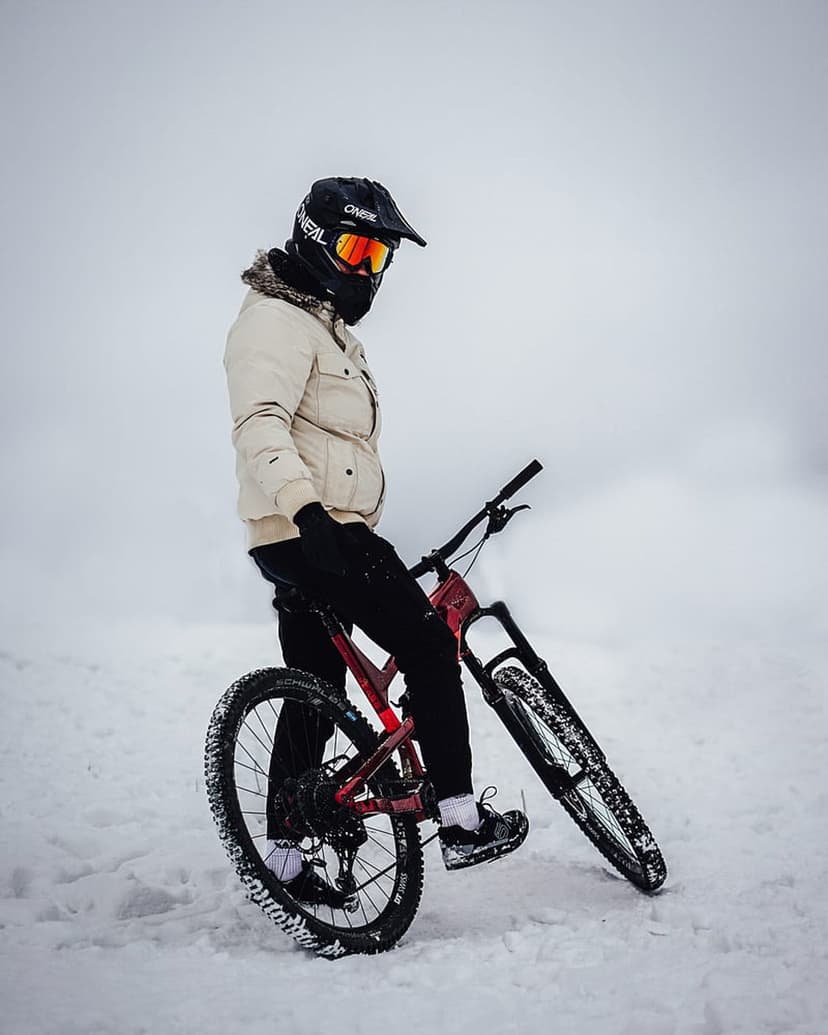 Mountain biker in a full-face helmet with a red bike stands on a snow-covered trail during a winter ride.