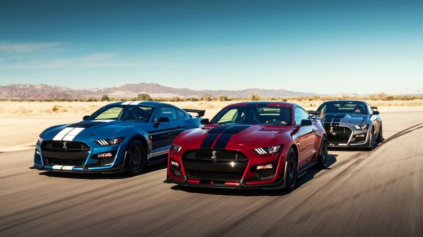 Three Ford Mustang Shelby GT500 cars in blue, red, and silver driving fast on a desert road under a clear sky.