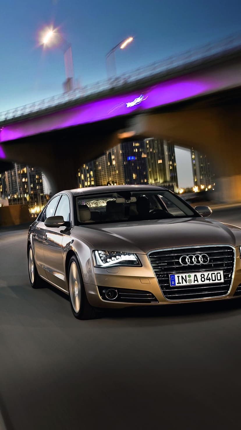 Gold Audi A8 sedan with glowing headlights on a city street at night near a purple neon suspension bridge.
