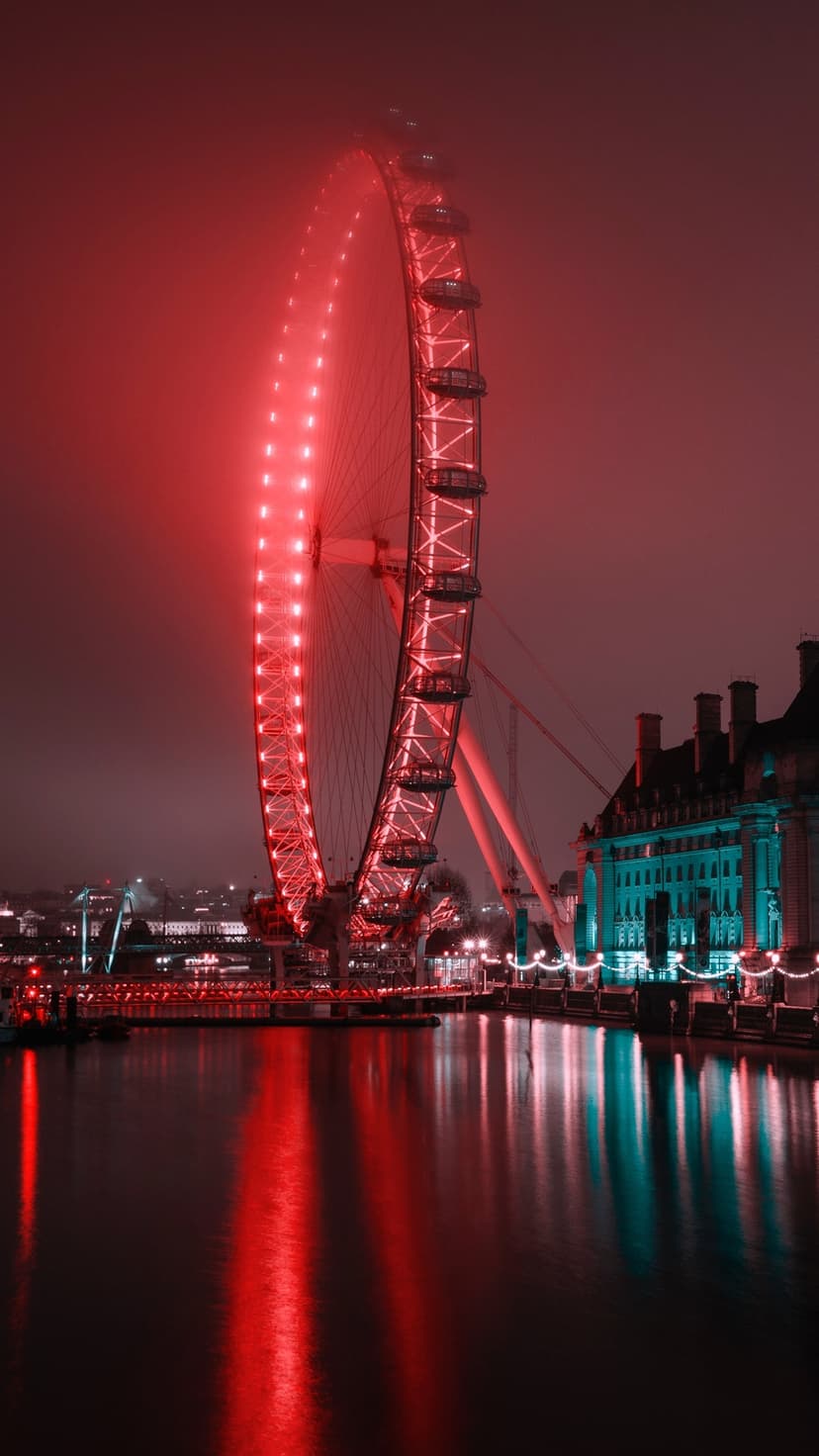 London Eye glowing bright red through thick fog with reflections on dark river water and distant teal lights.