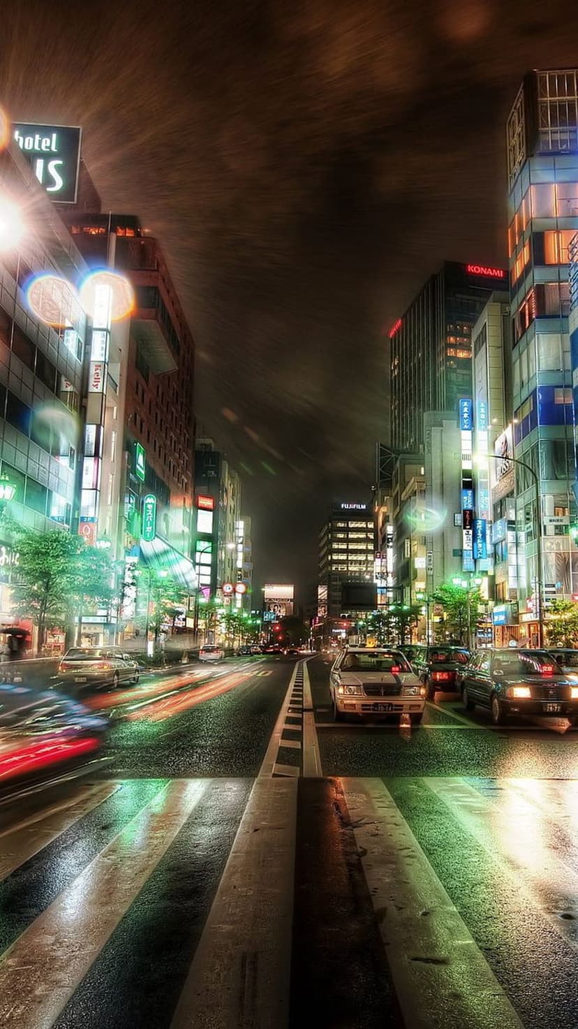 Red and white vehicle light trails stretch across a rain-slicked Tokyo street surrounded by glowing neon signs.
