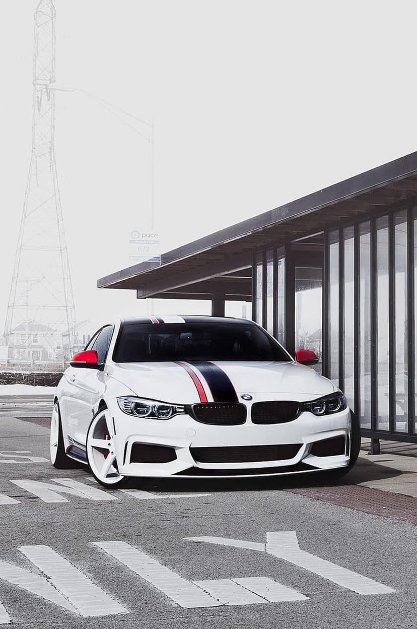 White BMW M4 coupe with red racing stripes parked before a glass building and an industrial transmission tower.