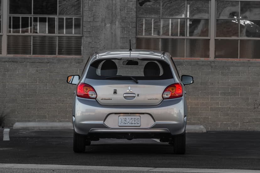 Rear view of a silver Mitsubishi Mirage parked in front of a grey brick building with industrial windows.