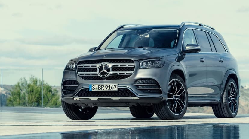 A grey Mercedes-Benz GLS luxury SUV parked on a reflective modern surface against a soft overcast sky.