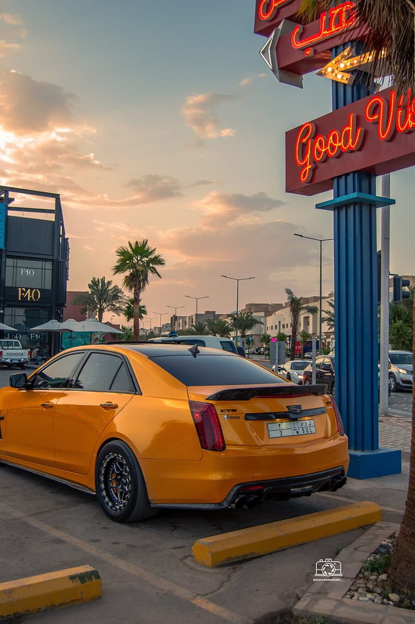 Orange Cadillac CTS-V sedan parked near a Good Vibes neon sign under a sunset sky with palm tree silhouettes.