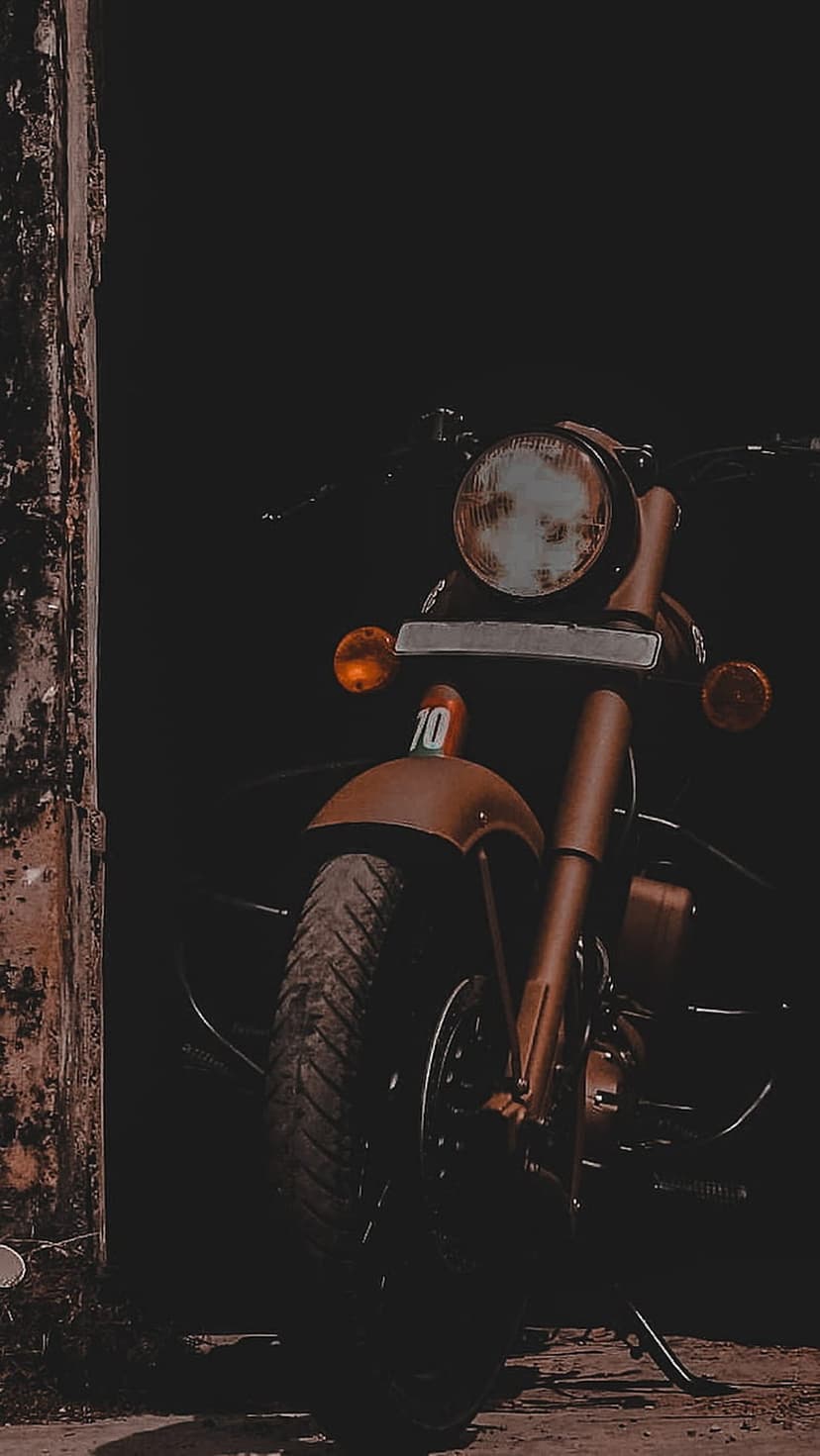 Vintage motorcycle headlight and orange turn signal emerge from dark shadows next to a textured wooden post.