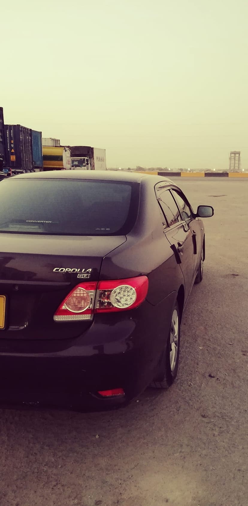 Dark Toyota Corolla GLi rear view in a shipping yard with stacked containers under an overcast, hazy sky.