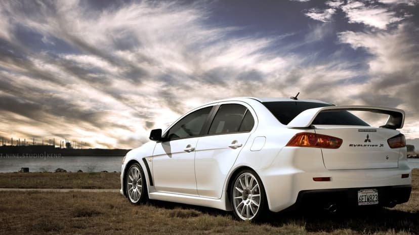 White Mitsubishi Lancer Evolution X with a large rear wing parked on grass against a dramatic cloudy sky.