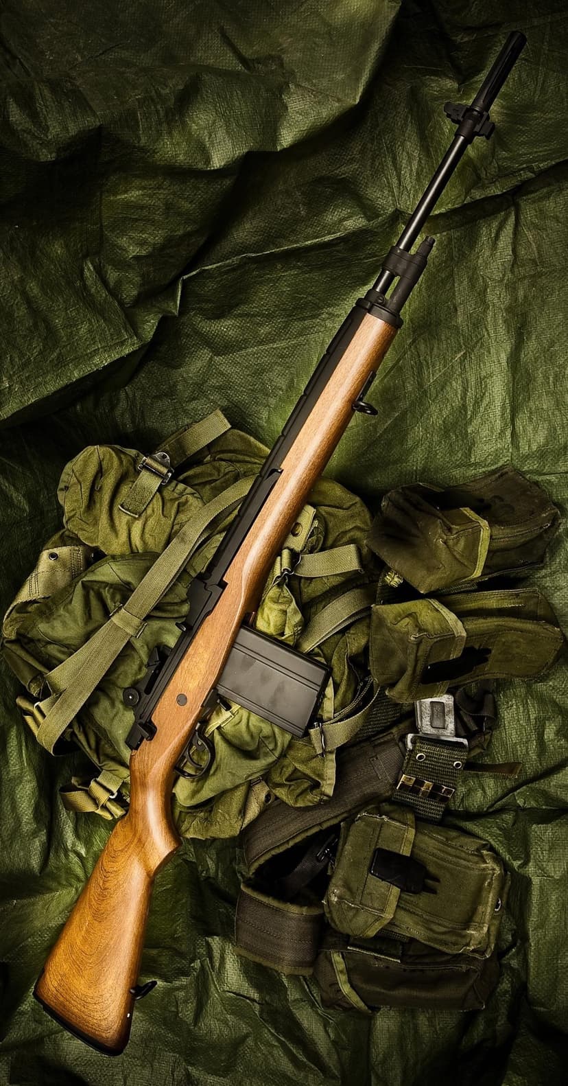 M14 rifle with a wooden stock rests on olive drab military gear and a green tarp in a vertical layout.