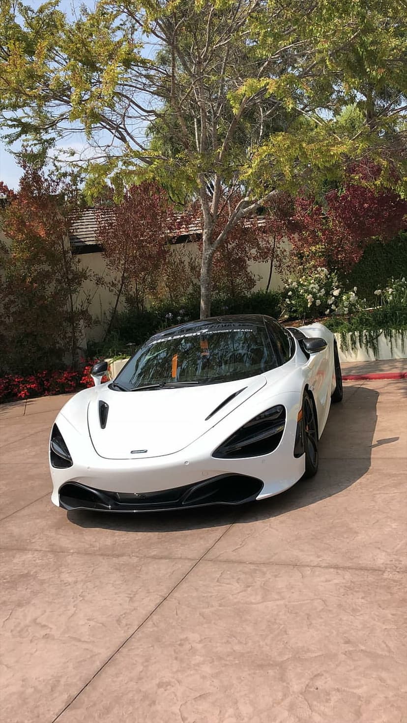 McLaren 720S in white parked on a driveway framed by red maple leaves and blooming white flowers on a sunny day.