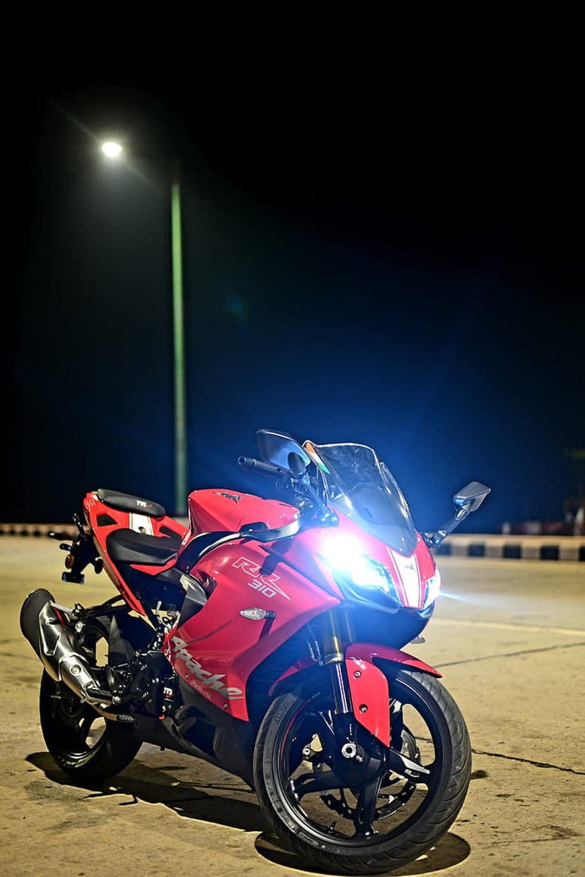Red TVS Apache RR 310 motorcycle with glowing headlights parked on a dark street under a yellow street lamp.