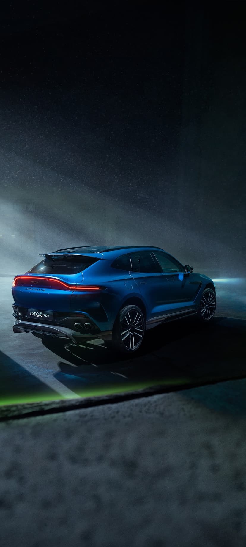 Blue Aston Martin DBX 707 SUV with glowing red taillights parked in a dark misty environment under cinematic light.