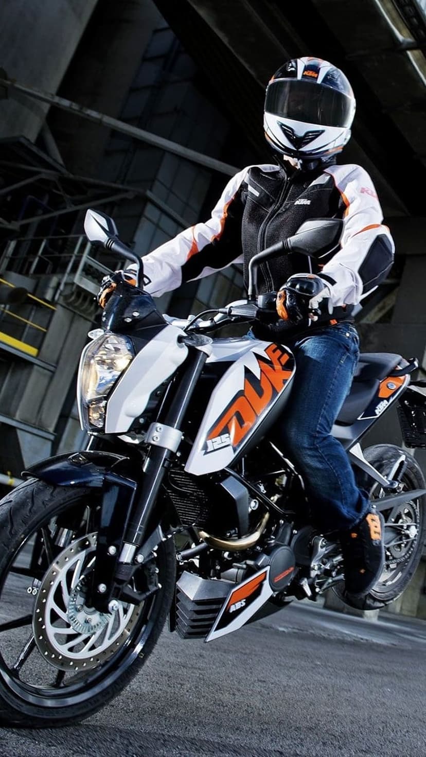 KTM rider in protective gear on a white and orange sportbike against a dark industrial warehouse background.