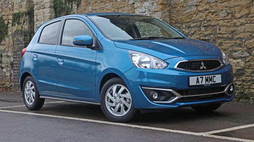 Blue Mitsubishi Mirage hatchback parked on wet pavement beside a rustic stone wall in soft daylight.