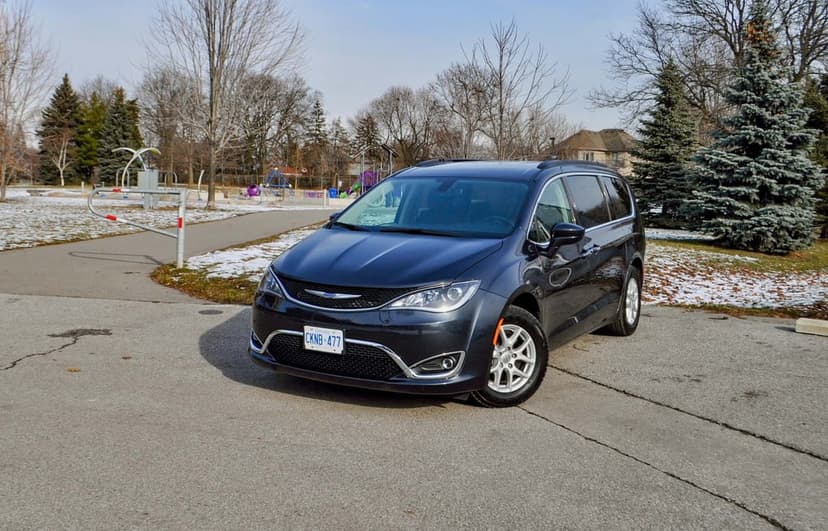 Dark charcoal Chrysler Pacifica minivan parked on pavement in a suburban winter park with light snow and bare trees.