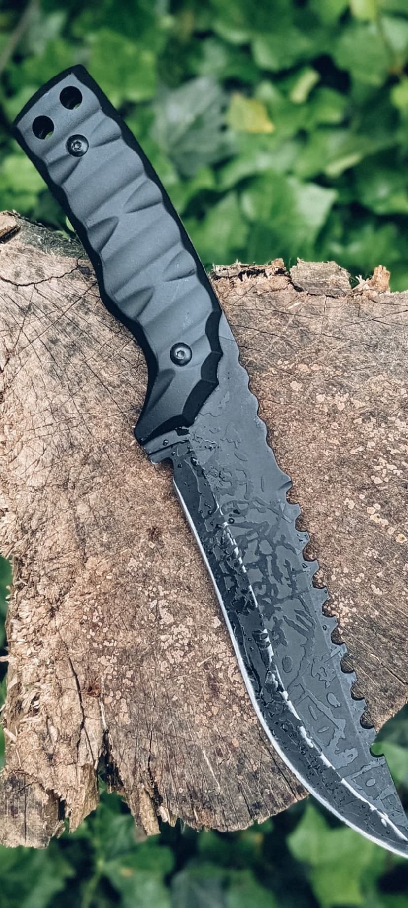 Tactical serrated knife with a gray handle lies on a weathered log against a backdrop of soft green foliage.