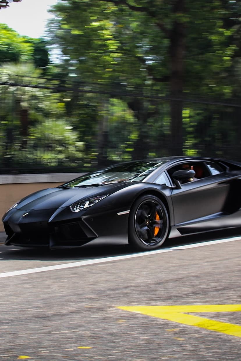 Matte black Lamborghini Aventador with orange calipers speeding past green trees on a paved race track.