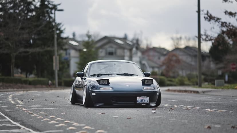 Navy blue stanced Mazda Miata on a grey street featuring extreme wheel camber and sleepy eye headlights.
