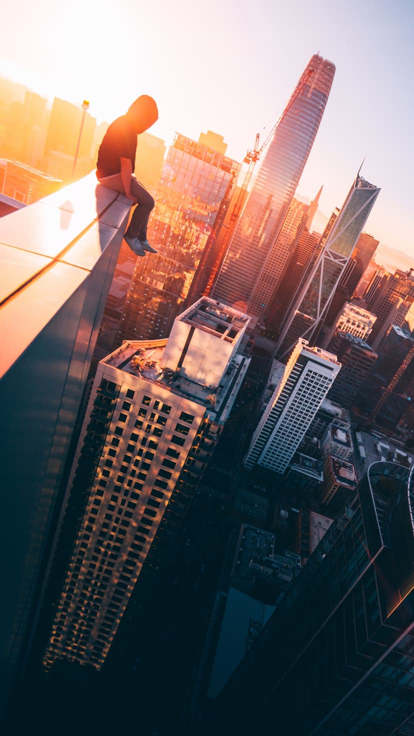 Person sitting on a skyscraper ledge above a sunlit city skyline with golden light reflecting off buildings.