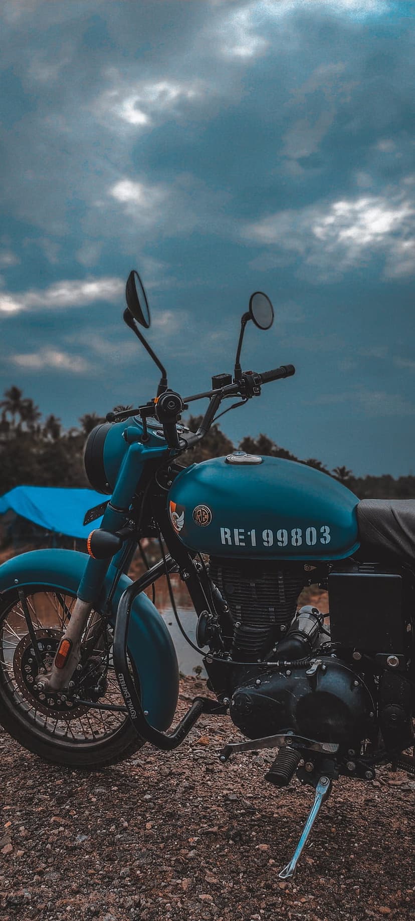 Blue Royal Enfield motorcycle with a vintage fuel tank stands before a dramatic overcast sky and lake scenery.